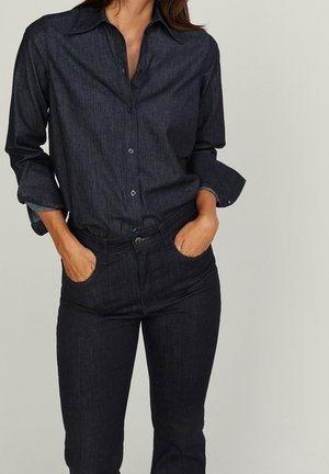 Woman wearing a dark denim button-up shirt with rolled sleeves and matching high-waisted jeans, hands in front pockets against a light background.