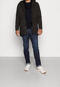 Mörkgrön parkas med huva, dragkedja framtill och dragsnören. Bärs över en svart tröja och kombineras med mörka jeans och vita sneakers.