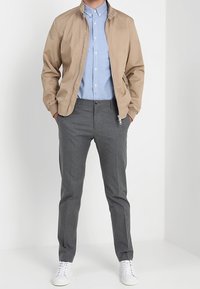 Giacca bomber beige, camicia azzurro chiaro con bottoni, pantaloni grigi su misura e sneakers bianche. Tessuti lisci e elementi di design semplici.