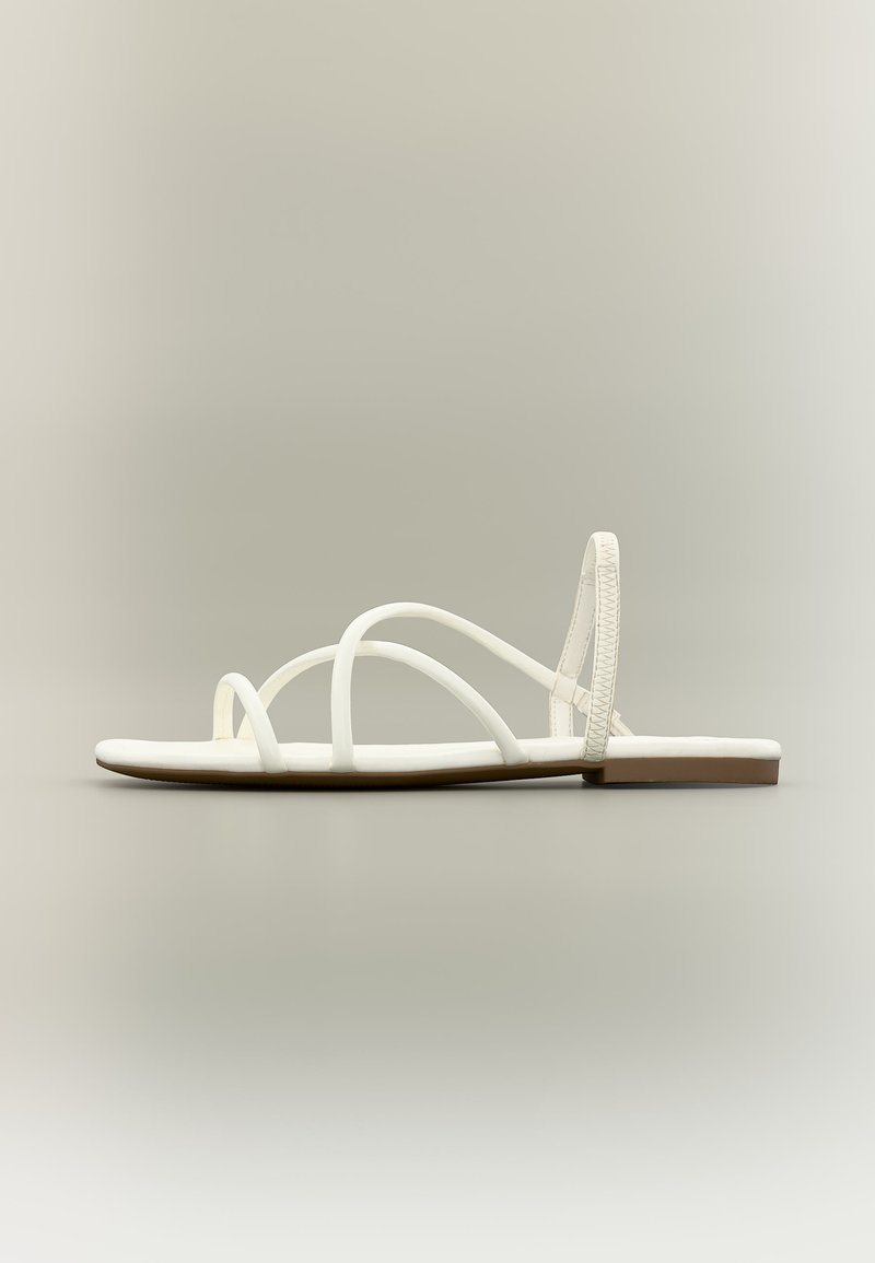 Λευκό επίπεδο σανδάλι με λεπτά διασταυρούμενα λουριά και λουρί στον αστράγαλο, παρουσιασμένο σε ουδέτερο φόντο.