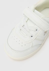 Sneakers blancs pour tout-petit avec une pointe perforée, des lacets et une sangle velcro sur un fond blanc.
