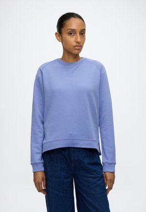 Donna con capelli scuri raccolti in uno chignon, indossa una felpa lavanda e jeans blu scuro, in piedi davanti a uno sfondo bianco semplice.