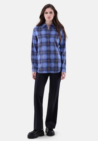 Blaue karierte Hemd mit langen Ärmeln, das eine Mischung aus hellblauen und dunkelblauen Farben mit braunen Akzenten zeigt. Kombiniert mit schwarzen geraden Hosen.