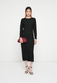 Vestuário longo preto de manga comprida com riscas texturizadas, até à metade da panturrilha. Acompanhado por uma carteira bordeaux e sapatos de salto pretos com tiras. Design simples.