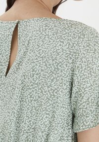 Haut vert à manches courtes présentant un motif abstrait texturé et éparse, avec un détail à trous dans le dos maintenu par un petit bouton.