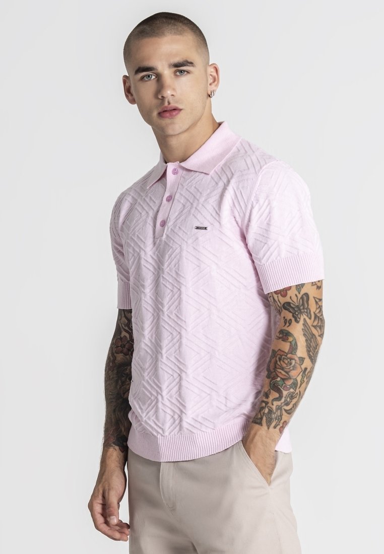 Hombre con cabello corto y tatuajes en ambos brazos, vistiendo una camisa polo de manga corta texturizada de color rosa claro y pantalones beige, con las manos en los bolsillos.