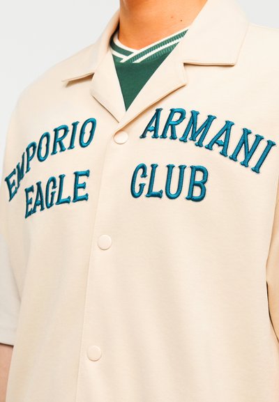 Μπεζ πουκάμισο με κουμπιά και κεντημένο κείμενο σε τιρκουάζ χρώμα "EMPORIO EAGLE" και "ARMANI CLUB", φορεμένο πάνω από πράσινο ριγέ μπλουζάκι με στρογγυλή λαιμόκοψη.