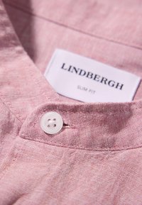 Colletto di camicia in lino rosa con una texture liscia, dotato di un pulsante bianco e di un'etichetta del marchio che recita 'LINDBERGH SLIM FIT.'