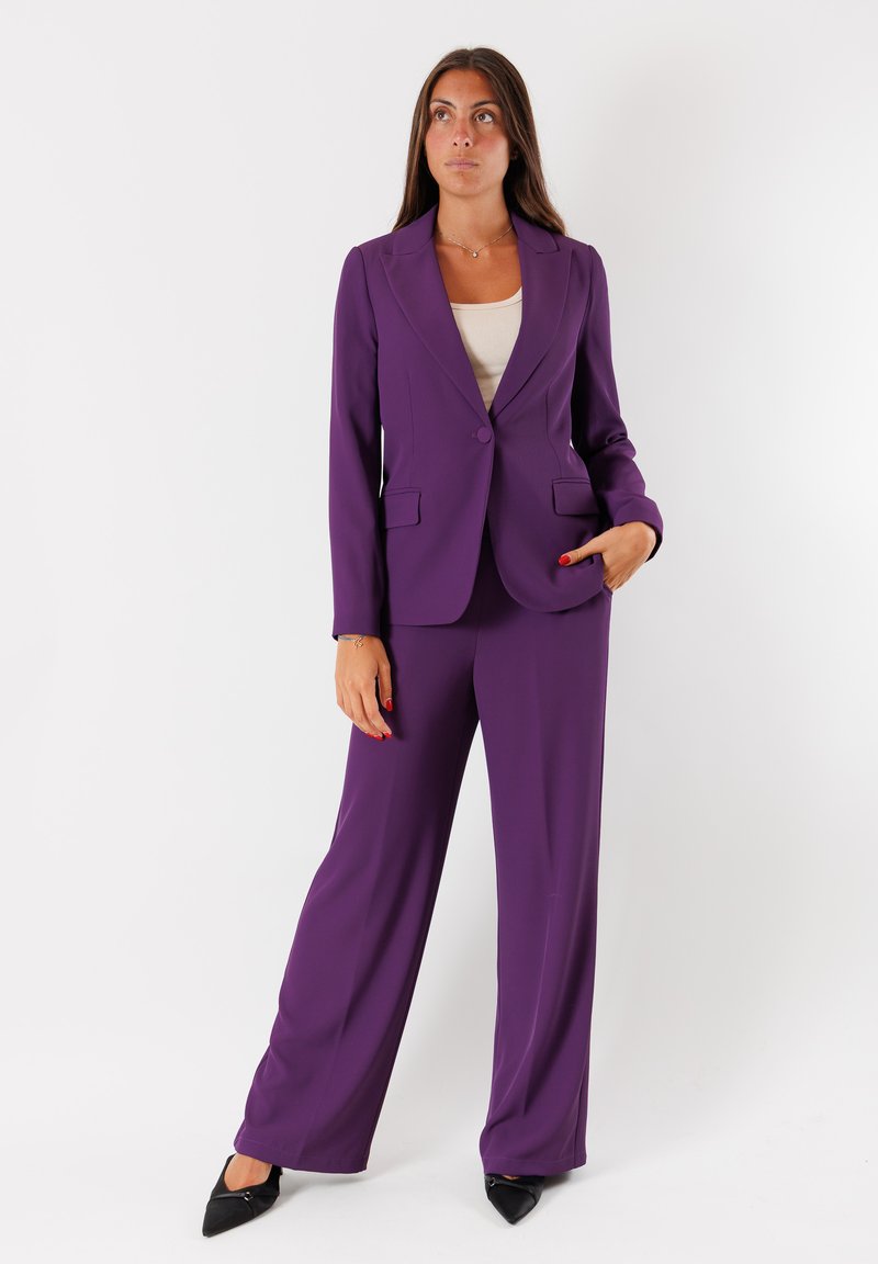 Blazer viola su misura e pantaloni a gamba larga, realizzati in tessuto liscio, con un bottone singolo, revers a punta e tasche frontali. Tacchi neri.