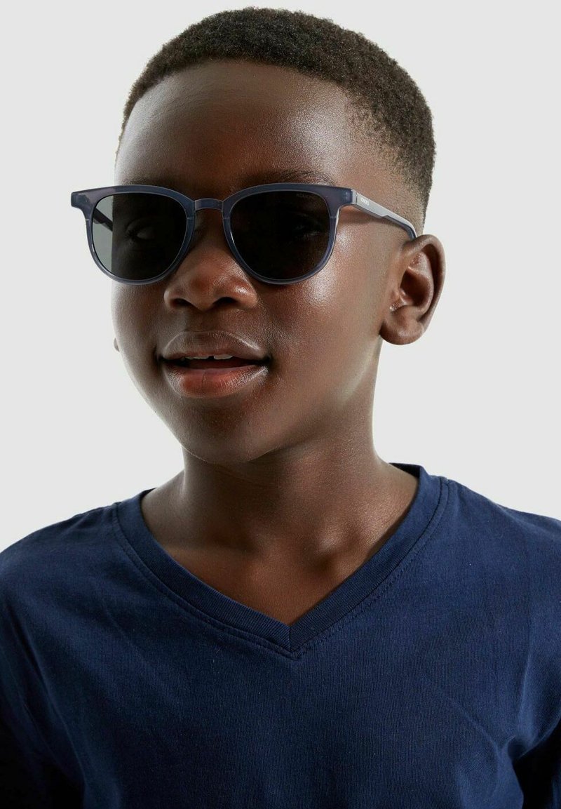 Lunettes de soleil noires avec une monture ronde, des verres foncés et un bandeau bleu marine. Portées par un enfant en t-shirt bleu marine à col en V. Teint clair.