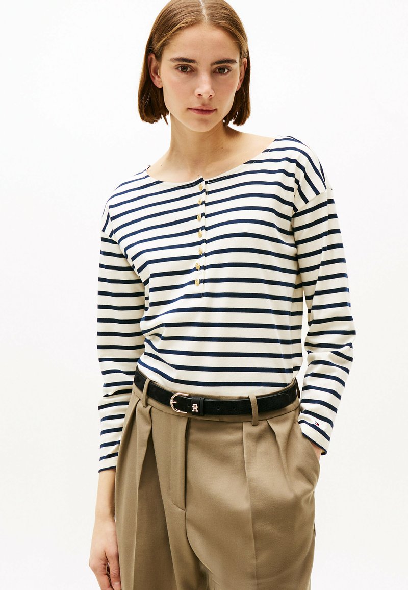 Camicia a righe a maniche lunghe in crema e blu navy, con scollatura a barca e dettagli con bottoni dorati. Abbinata a pantaloni plissettati beige e una cintura nera.