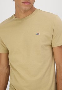 Tricou din bumbac bej, cu guler rotund, mâneci scurte și un mic emblem cu dungi roșii, albe și albastre pe pieptul stâng.