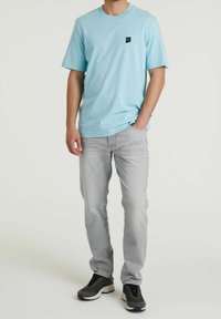 Jasnoniebieski T-shirt z krótkim rękawem z czarną łatką logo, w połączeniu z szarymi dżinsami i czarnymi butami sportowymi. Luźny krój i lekki materiał.