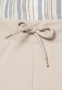 Beige cotton joggers with a textured waistband and a drawstring tie. The tie features gold metal tips. Vertical blue and white striped fabric above.