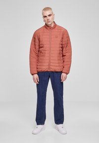 Polstret jakke i rustfarge med høy krage, horisontalt quiltet mønster, glidelås foran og sidelommer, sammen med blå jeans og hvite joggesko.