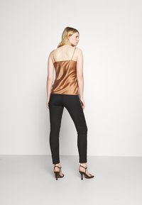 Camisola in satin marrone con spalline sottili, abbinata a pantaloni neri attillati e tacchi neri con cinturini. La modella è mostrata di spalle.