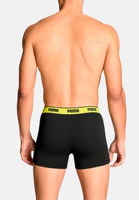 Schwarze Boxershorts mit einem gelben Puma-Logo-Bund, die eng anliegen und aus weichem Material bestehen, von hinten vor einem weißen Hintergrund betrachtet.