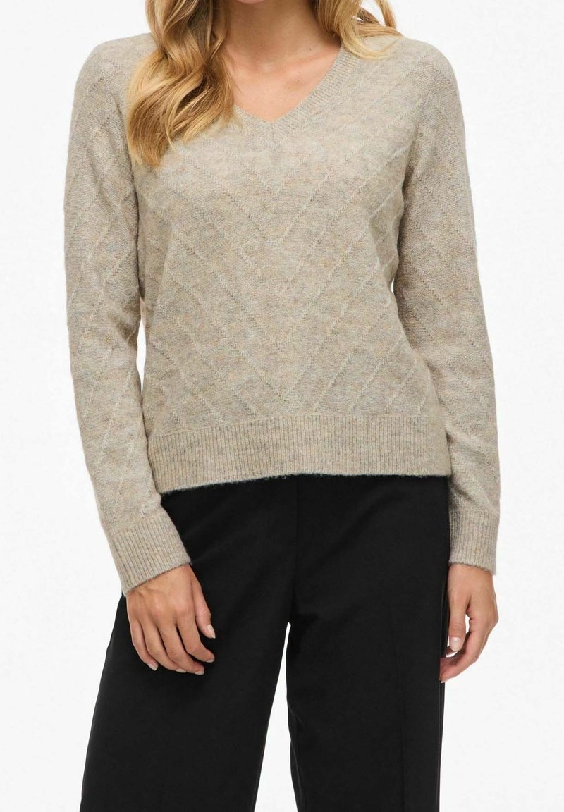 Femme portant un pull beige à col en V en maille torsadée à manches longues et un pantalon noir, debout devant un fond uni.