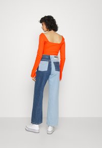 Camisola laranja de mangas longas com um design quadrado nas costas, combinada com jeans de ganga de pernas largas em azul escuro e claro. Ténis brancos.