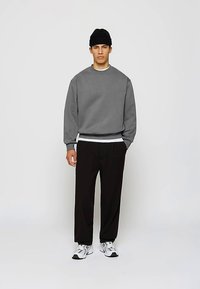 Homme mannequin portant un sweat-shirt gris, un bonnet noir, un pantalon noir et des baskets blanches, se tenant devant un fond gris clair uni.