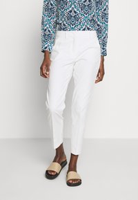 Osoba w białych, dopasowanych spodniach, długim rękawie w niebiesko-białe wzory oraz beżowych klapkach na czarnych podeszwach.