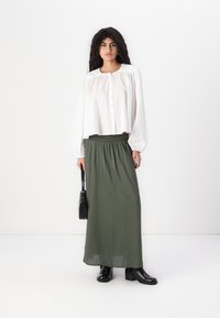 Blouse blanche à manches longues avec un boutonnage à l'avant, associée à une jupe maxi fluide verte. La tenue est complétée par des bottines noires et un petit sac à main.