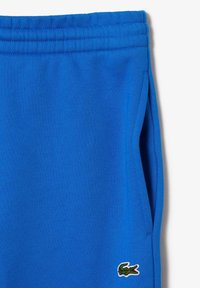 Blå bomullsoveraller med elastisk midjeband, sidofickor och en liten broderad krokodillogotyp på nederbenet.