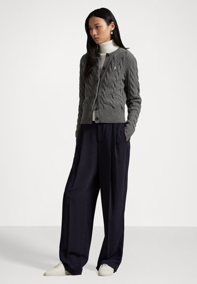 Grijze kabelgebreide cardigan met een ronde hals en knoopsluiting, gedragen over een witte coltrui. Gecombineerd met brede zwarte broek en crèmekleurige schoenen.