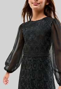 Robe en dentelle noire avec un corsage texturé orné de motifs floraux, des manches longues transparentes et un détail de taille cintrée.