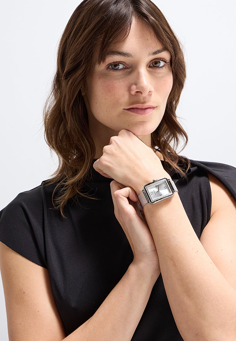 Mujer con cabello castaño hasta los hombros, vestida con una blusa negra y un reloj de pulsera rectangular de plata en la muñeca izquierda.