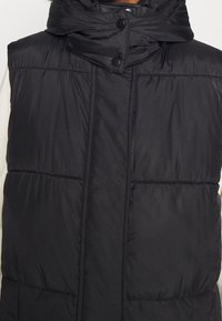 Gilet noir matelassé sans manches avec col montant et fermeture à boutons-pression, porté par-dessus un vêtement à manches longues de couleur claire.