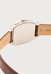 Πίσω μέρος ασημένιου ρολογιού Tommy Hilfiger με καφέ δερμάτινο λουράκι και δύο πλευρικά κουμπιά σε λευκό φόντο.