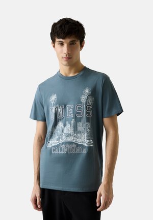 Joven con una camiseta azul con el paisaje urbano blanco de Los Ángeles, palmeras y el texto "Guess California", de pie contra un fondo liso.