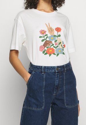 Wit T-shirt met afbeelding van drie diverse handen tussen kleurrijke bloemen, gecombineerd met een high-waisted blauwe spijkerbroek met voorzakken.