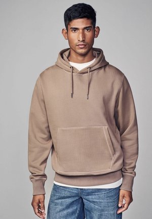 Brauner Kapuzenpullover mit einer Vorderseite-Tasche, gerippten Bündchen und Kordelzug. Wird über einem weißen T-Shirt getragen und mit blauen Jeans kombiniert.