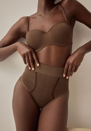 Nő barna pánt nélküli melltartóban és magas derekú alakformáló fehérneműben, kezét csípőjén tartva pózol semleges háttér előtt.
