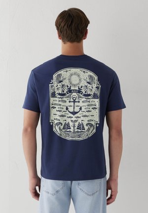 Mann trägt dunkelblaues T-Shirt mit großem nautischem Design auf dem Rücken, das Sonne, Anker, Fisch, Vögel, Palmen und Segelboote zeigt.