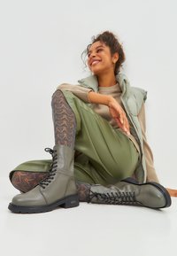 Olijfgroene veterschoenen met een glad leren afwerking, zwarte rubberen zool en leggings met een gemêleerd patroon in tinten bruin en zwart.