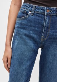 Mėlynos džinsinės kelnės aukštu liemeniu, turinčios penkias kišenes, vario spalvos sagos užsegimą ir kontrastingas oranžines siūles ant lygios tekstūros.