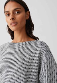 Femme aux cheveux châtains mi-longs portant un pull gris texturé avec des rayures noires diagonales, tournée légèrement vers la gauche devant un fond uni.