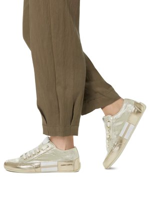 Sneaker dorate metalliche con dettagli bianchi, punta rotonda, upper texture, suola piatta e chiusura con lacci. Indossate con pantaloni marroni a vestibilità ampia.