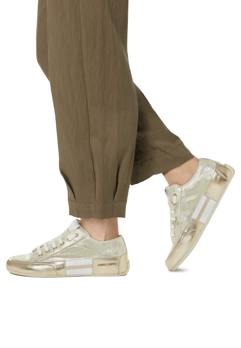 Sneaker dorate metalliche con dettagli bianchi, punta rotonda, upper texture, suola piatta e chiusura con lacci. Indossate con pantaloni marroni a vestibilità ampia.