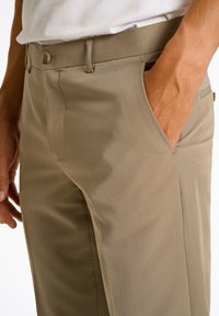 Beige Chinos mit glatter Textur, ausgestattet mit einem standardmäßigen Knopfverschluss und Seitentaschen. Taillierte Passform mit klassischem Design.