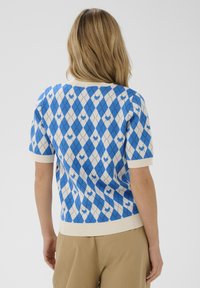 Kék-fehér rövid ujjú pulóver, amely gyémántmintával, bordázott gallérral és mandzsettákkal rendelkezik. Puha textúrájú, részletes kidolgozottsággal.