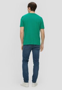 Man in een groen T-shirt met korte mouwen, blauwe jeans en witte sneakers, staand met zijn rug naar de camera op een effen lichte achtergrond.