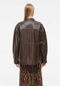 Veste en cuir brun avec une coupe décontractée, dotée d'un col de chemise et de manches longues. Le dos présente une texture lisse et brillante.
