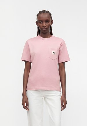 Kobieta z zaplecionymi włosami, ubrana w jasnoróżowy t-shirt z krótkim rękawem z logo na przedniej kieszeni oraz białe spodnie, stojąca na jednolitym tle.