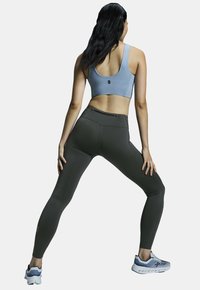 Soutien-gorge de sport bleu clair, leggings taille haute vert foncé avec poche zippée, associés à des chaussures de sport grises avec des accents bleus.