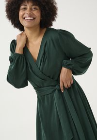 Abito avvolgente verde scuro con scollatura a V profonda, maniche lunghe a sbuffo e una cintura legata in vita. Tessuto morbido e fluido con dettagli plissettati.
