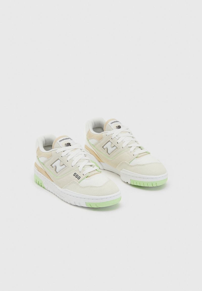 Paire de baskets New Balance 550 blanches et beiges avec des accents vert clair, des lacets blancs et des logos de marque sur un fond uni.